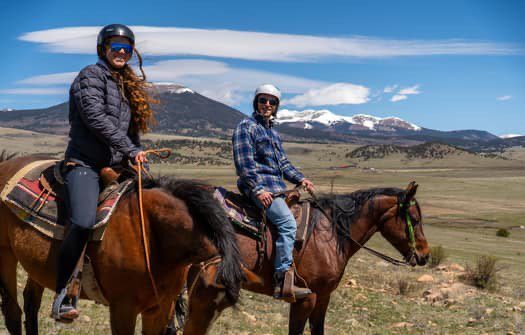 Two people horseback riding with mountain backdrop.